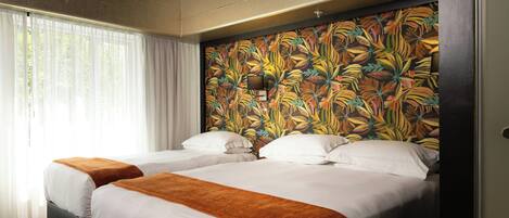 Habitación de hotel con dos camas, ropa blanca, cabecera con motivos de hojas tropicales y ventana con cortinas traslúcidas.