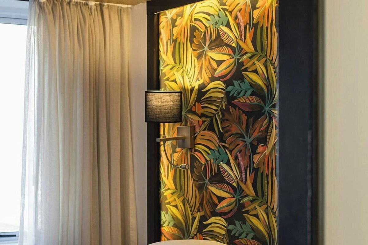 A hotel room with a bed, a window with curtains, and a wall with a tropical leaf pattern.