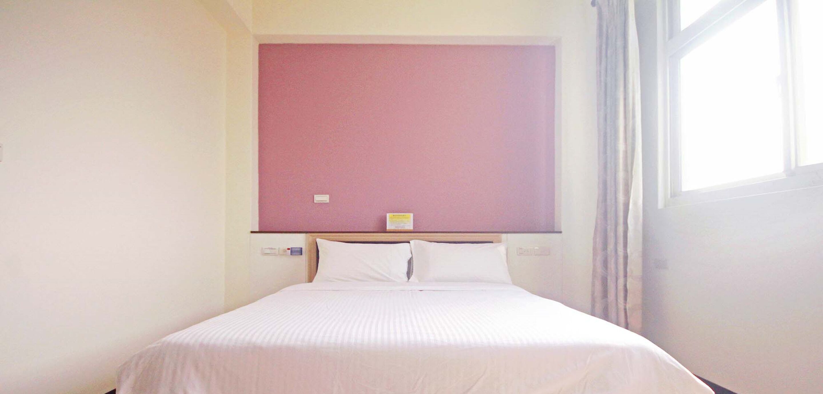 A hotel room with a single bed, a pink wall, and a window with curtains.