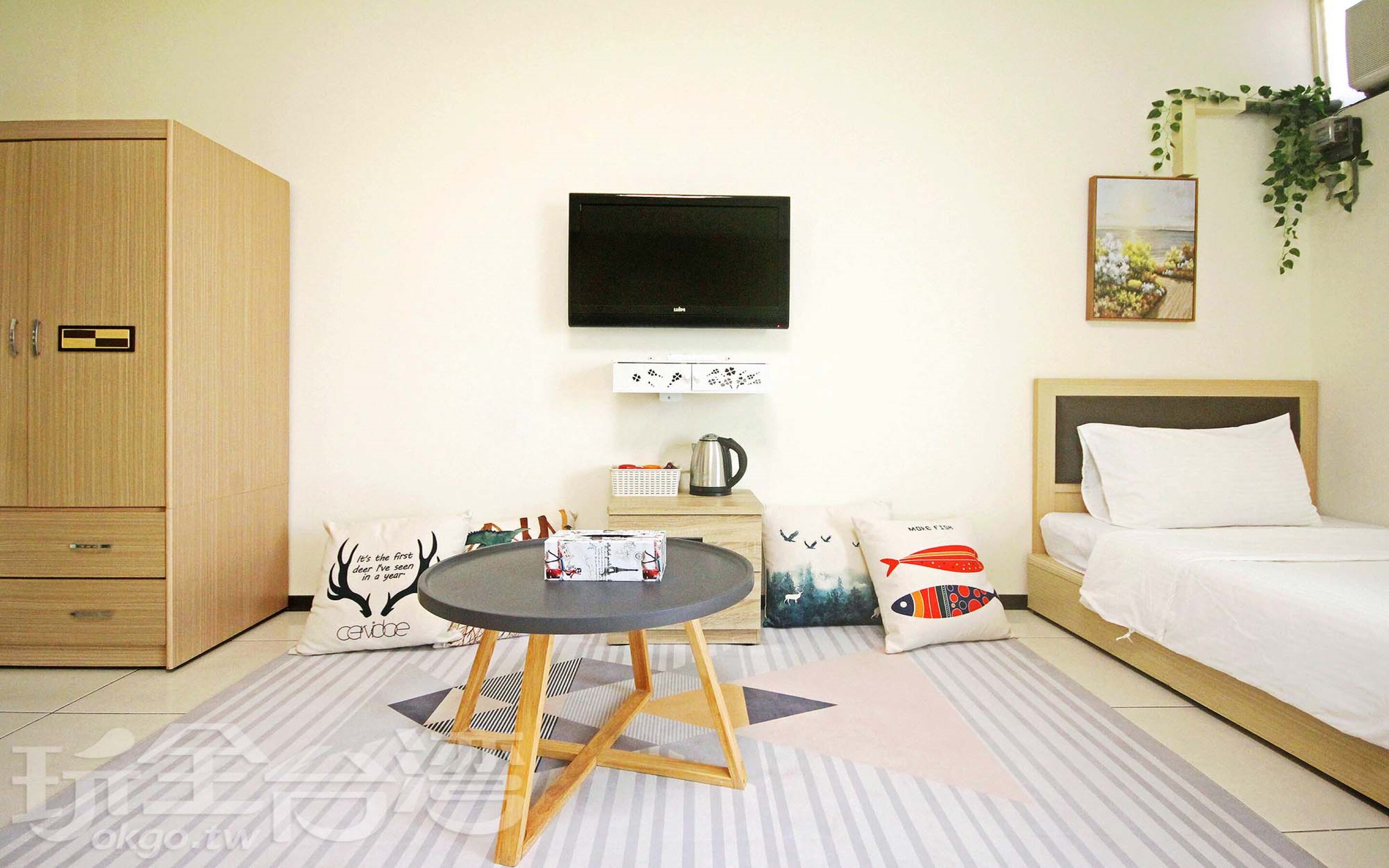 Una habitación de hotel con una cama, un televisor montado en la pared, un armario de madera, una mesita con una tetera y un cojín decorativo.