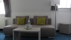 A modern hotel room with a grey sofa, two green pillows, a small white table with a bottle and glass, a floor lamp, and a window with curtains. - BSW Inselhotel Rote Erde Borkum (Borkum)