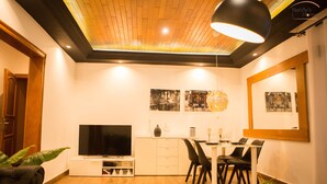 A modern living room with a flat-screen TV, dining table, chairs, and a large mirror.