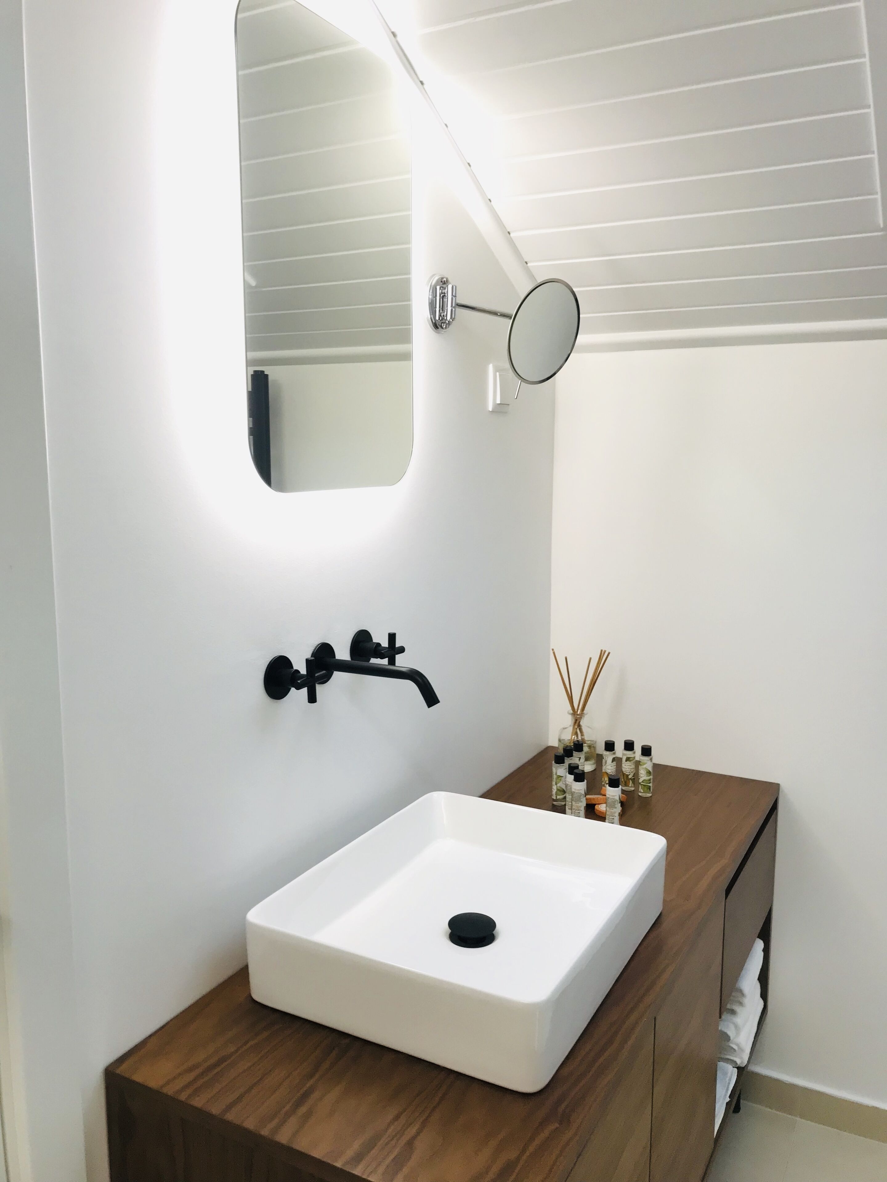 A bathroom with a white sink, black faucet, and mirror. There is a wooden vanity with a bottle of hand soap and reed diffusers.