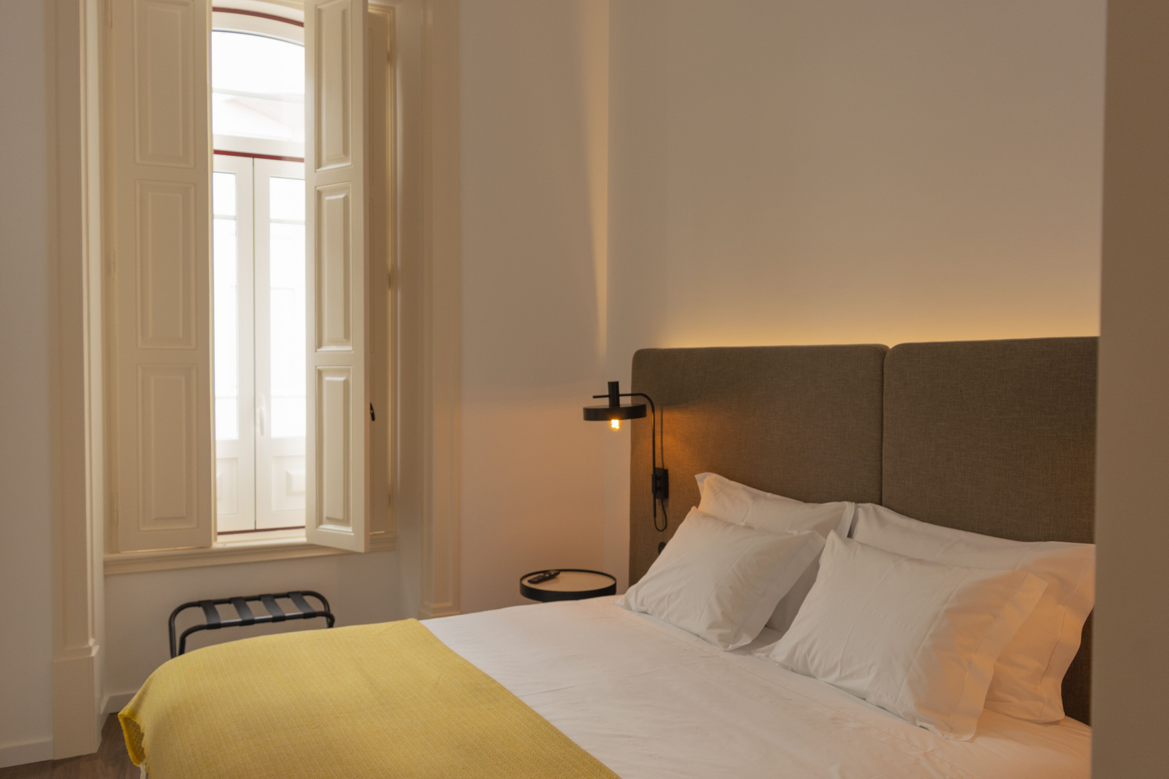 A neatly made bed with white linens and a yellow throw blanket, a bedside table with a lamp, and a window with a view of the outside.