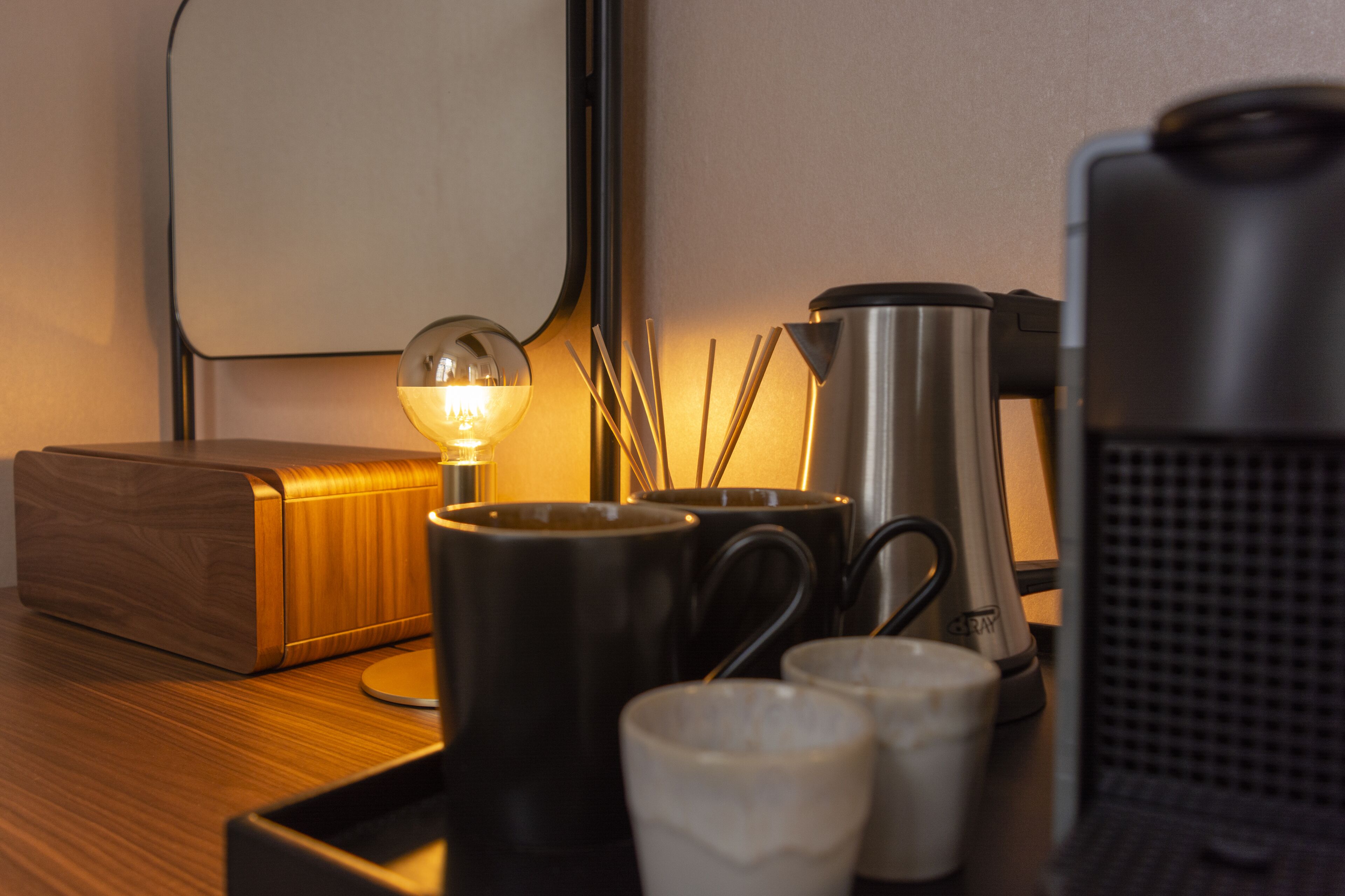 Uma mesa de cabeceira de madeira com uma luminária, duas canecas pretas, uma cafeteira de aço inoxidável e uma caixa de madeira.