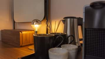 Uma mesa de cabeceira de madeira com uma luminária, duas canecas pretas, uma cafeteira de aço inoxidável e uma caixa de madeira.