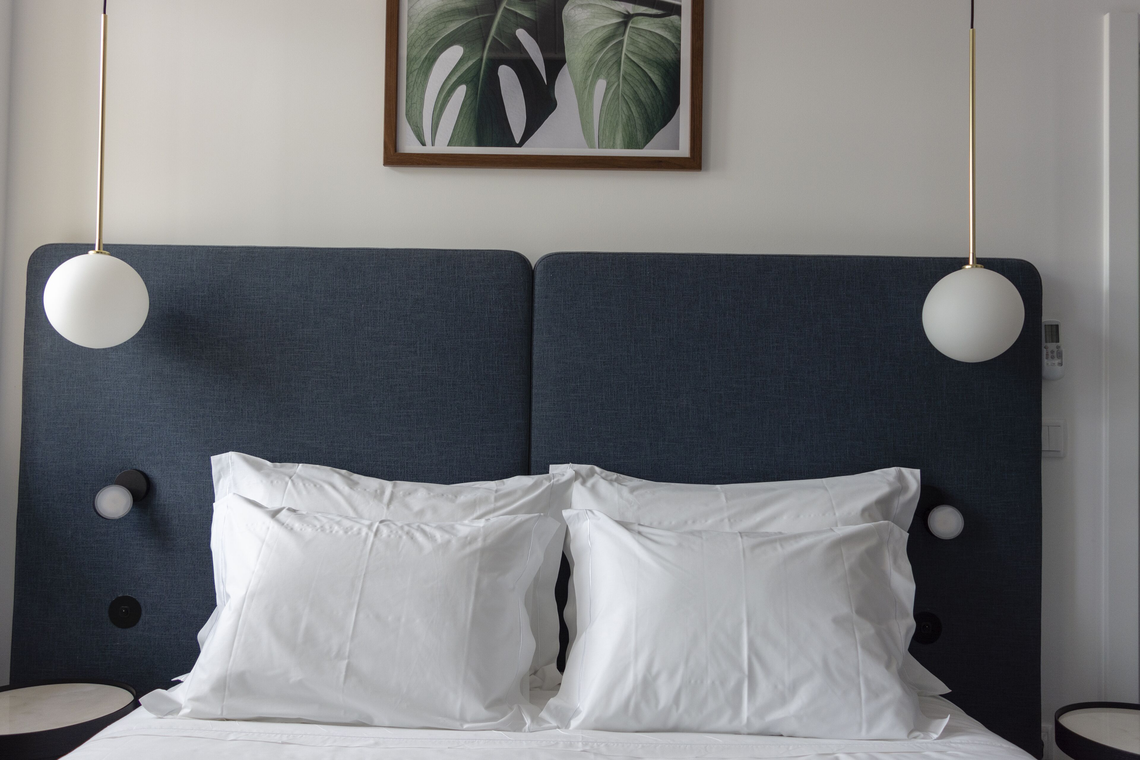 Una cama con ropa de cama blanca, cabecera azul oscuro, dos lámparas de noche y una planta enmarcada en la pared.