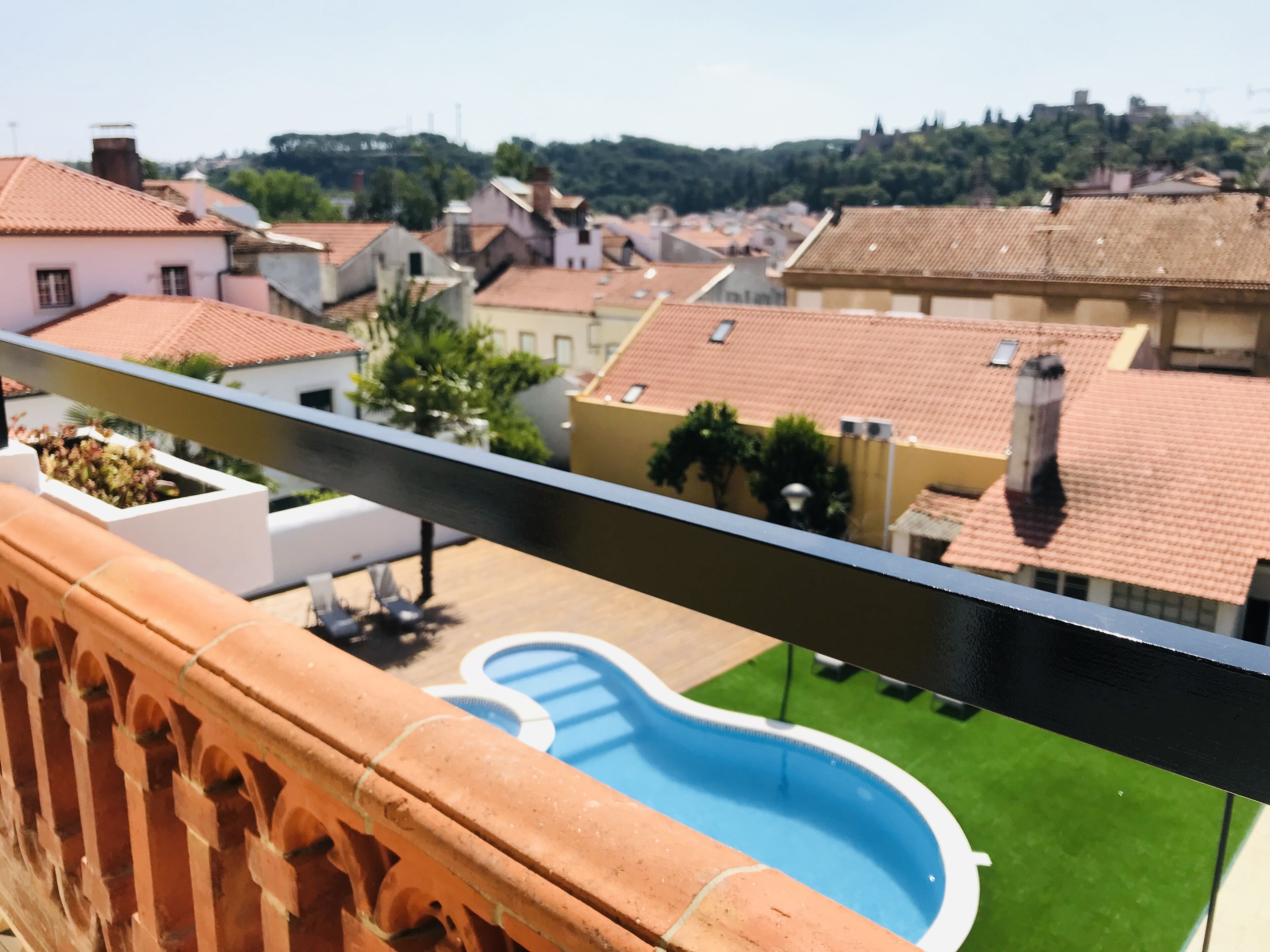 Un balcón con vista a una piscina, barandilla de terracota y casas con tejados de tejas rojas.