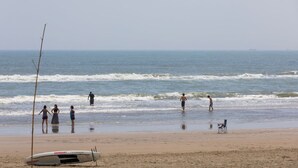 A beach with people walking and standing in the water, a lifeguard stand, and a surfboard on the sand. - Wave House 99 (Kujukuri)
