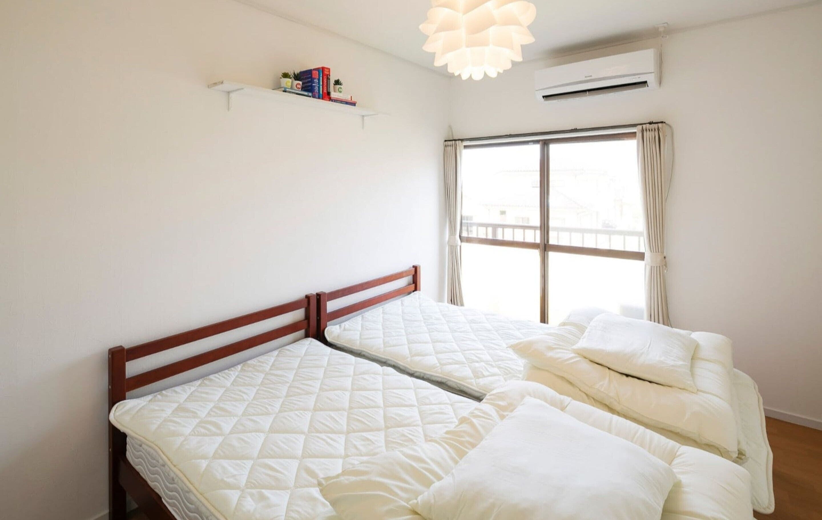 Un dormitorio con dos camas, una ventana con cortinas, una estantería con libros y un equipo de aire acondicionado.