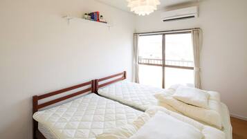 Un dormitorio con dos camas, una ventana con cortinas, una estantería con libros y un equipo de aire acondicionado.