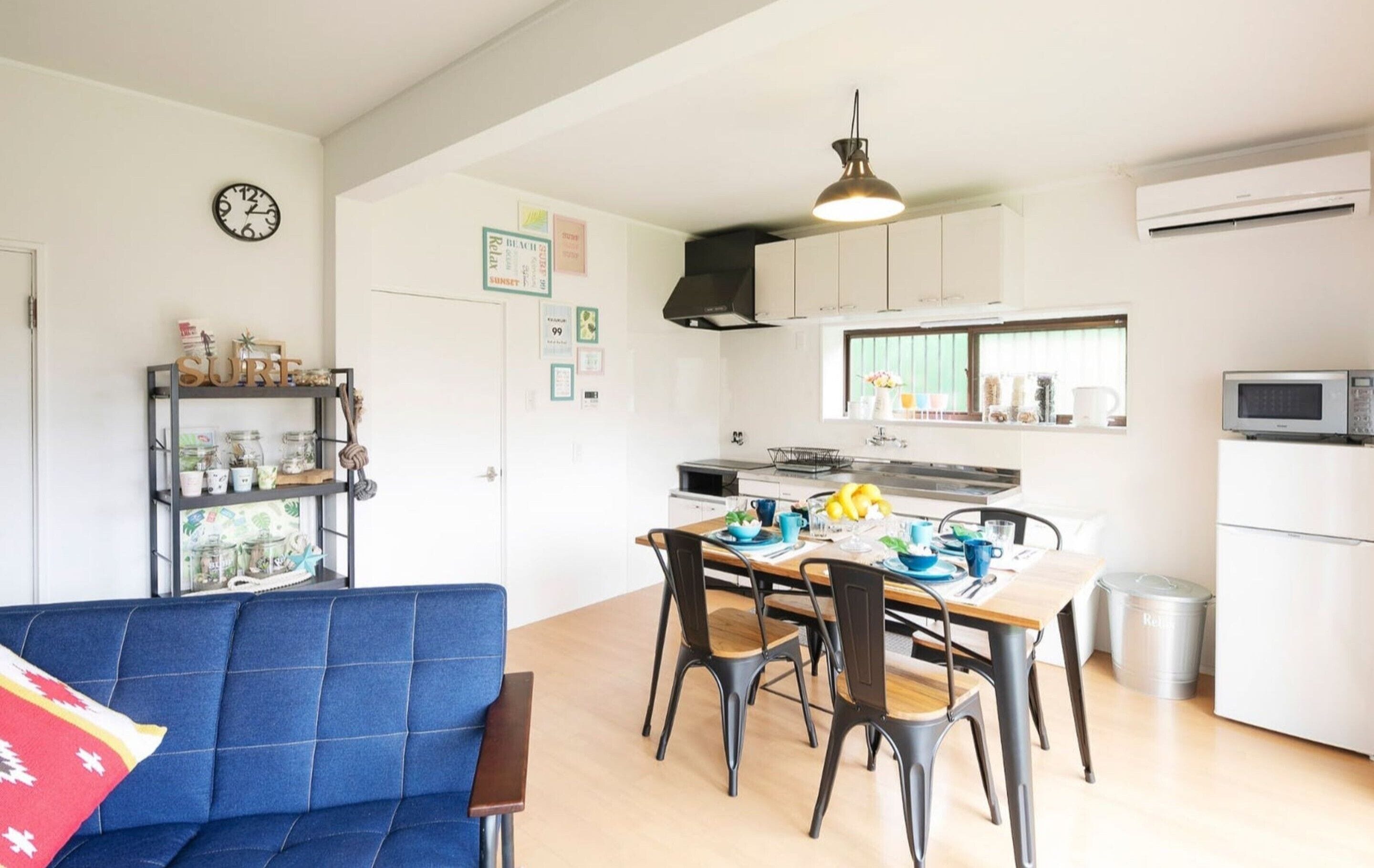 Een kleine keuken met witte kastjes, een eettafel met stoelen, een blauwe bank en een klok aan de muur.