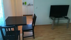 Uma área de refeições com mesa e cadeiras, uma cozinha ao fundo e uma TV em um suporte.