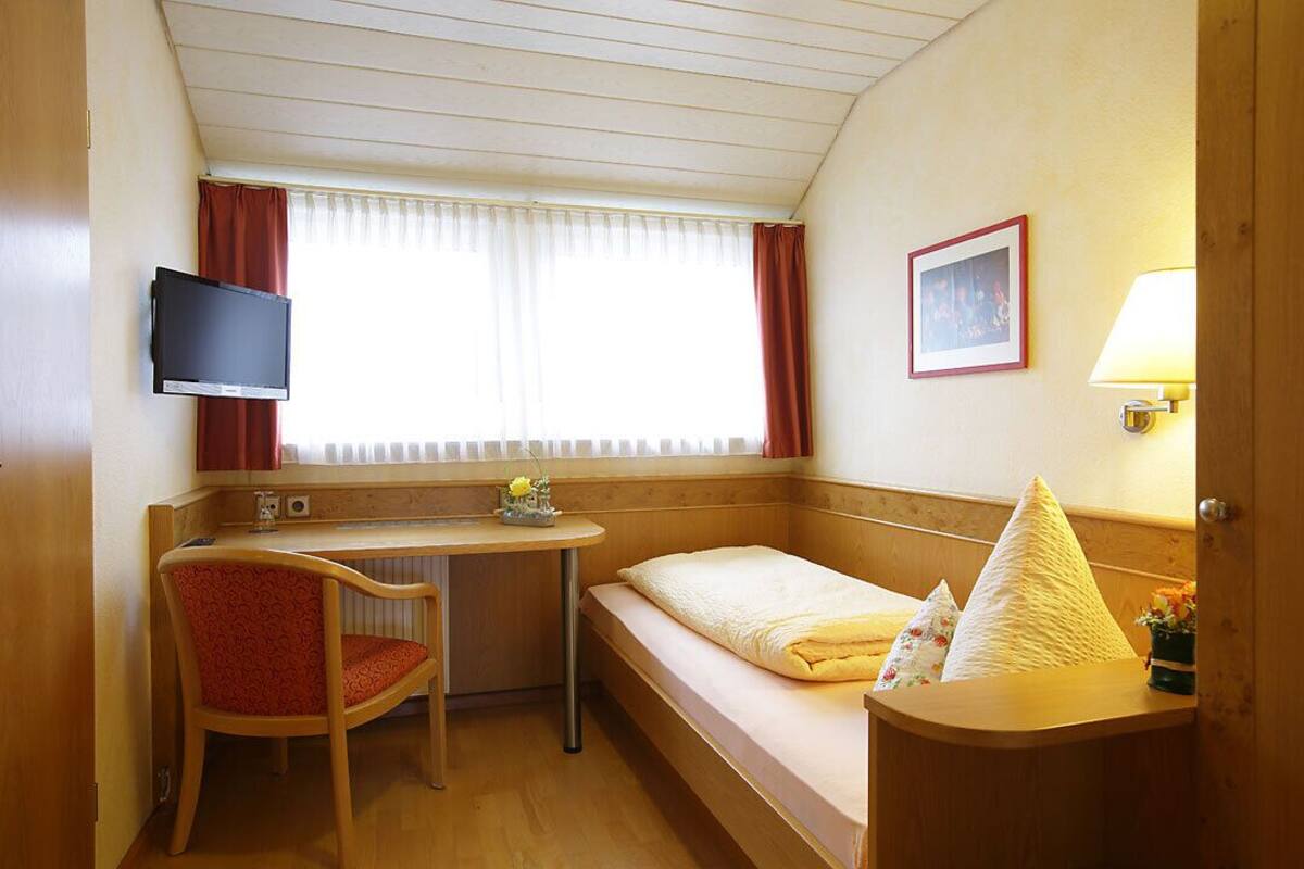 A small hotel room with a single bed, a desk with a chair, a television mounted on the wall, and a window with curtains.