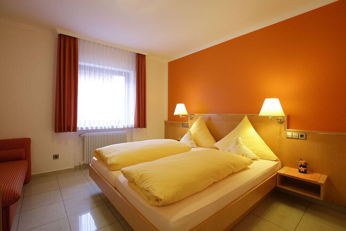 A hotel room with a double bed, two bedside lamps, a small bedside table, a red chair, and a window with curtains.