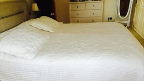 A neatly made bed with a white quilt, a wooden headboard, a bedside table with a lamp, and a dresser with drawers. - Lovely Family House in Sidcup (Sidcup)
