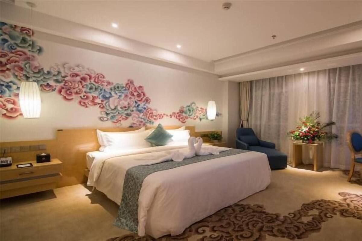 Habitación de hotel con una cama grande, un escritorio con silla, una mesita con planta y un mural decorativo en la pared.