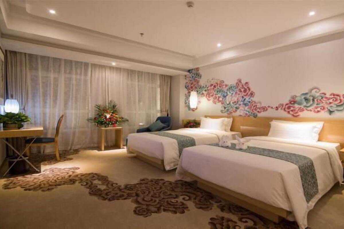 Habitación de hotel con dos camas, un escritorio con una silla, una mesita con una planta y un adorno en la pared.