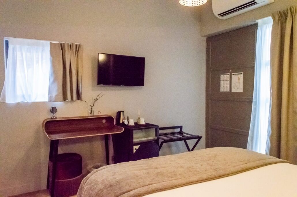 A hotel room with a bed, a desk with a chair, a flat-screen TV, and a window with curtains.