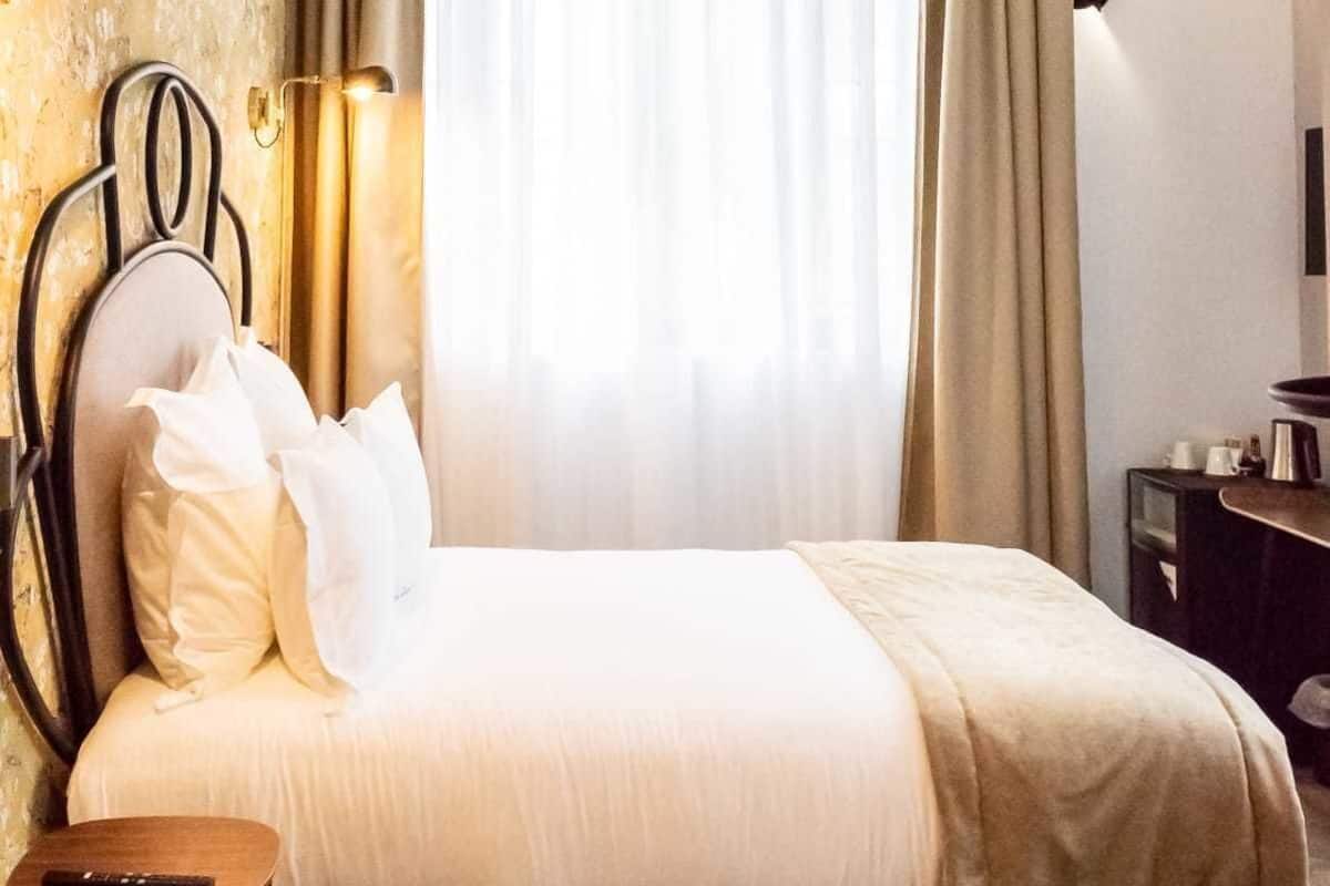 Une chambre d’hôtel comprenant un lit, une table de chevet, une lampe, des rideaux et un luminaire fixé au mur.