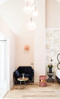 Una habitación moderna con un sillón azul oscuro, una mesita redonda pequeña, un reposapiés rosa y una mesita auxiliar negra con una planta.
