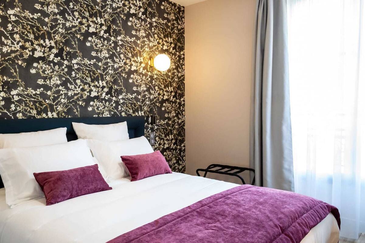 Une chambre d’hôtel avec un lit, du linge de lit blanc, un plaid violet, un papier peint à motifs floraux, une lampe fixée au mur et une fenêtre avec des rideaux.
