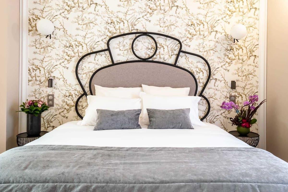 Un lit avec une tête de lit grise, une literie blanche et deux oreillers gris. De chaque côté du lit, on trouve deux tables de chevet sur lesquelles sont posées des lampes et des vases de fleurs.