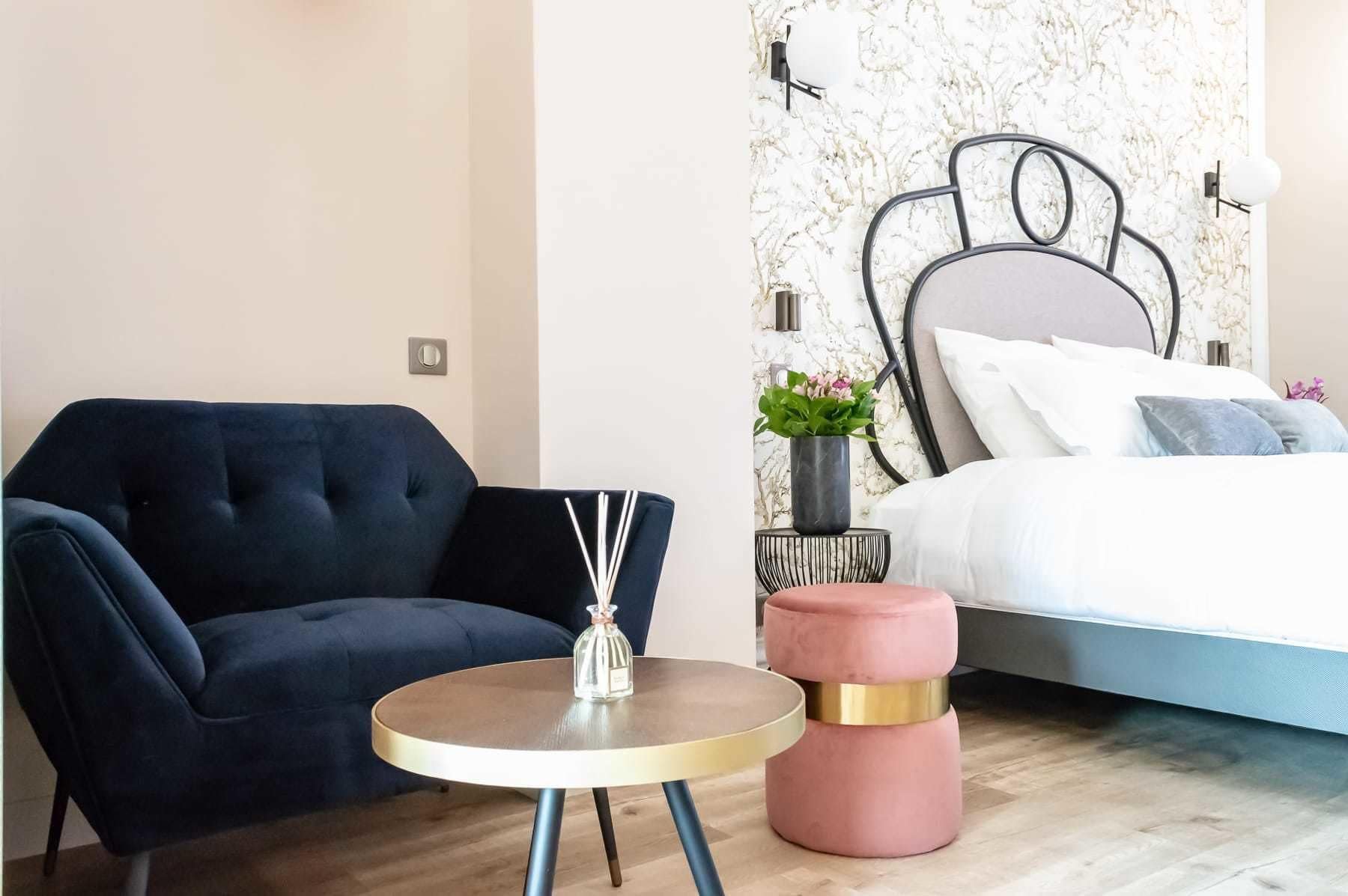 A bedroom with a bed, a round table with a glass vase, a blue armchair, and a pink ottoman.