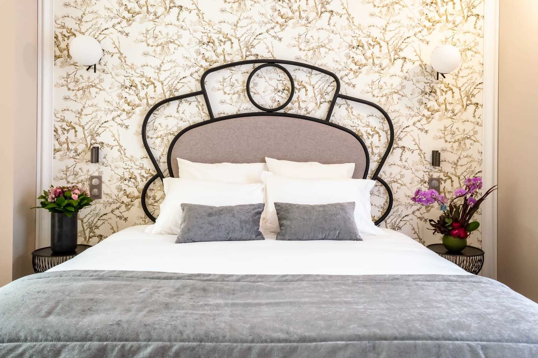 A neatly made bed with a grey duvet and pillows, a marble-patterned wall, and two bedside tables with lamps and flowers.