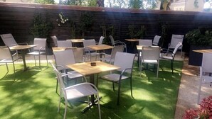 Un espace extérieur aménagé en terrasse, avec plusieurs tables et chaises, entouré d’une clôture en bois et de verdure.