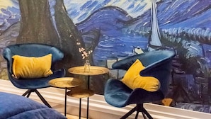 Une pièce avec un grand tableau représentant une scène nocturne, deux fauteuils bleus avec des coussins jaunes, une petite table ronde et un lit avec une tête de lit bleue.