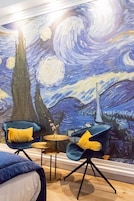 Habitación con un cuadro grande de una escena nocturna, dos sillones azules con cojines amarillos, una mesita redonda y una cama con cabecera azul.