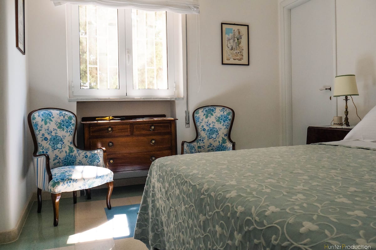 A bedroom with a bed, two chairs, a dresser, a window, and a painting on the wall.