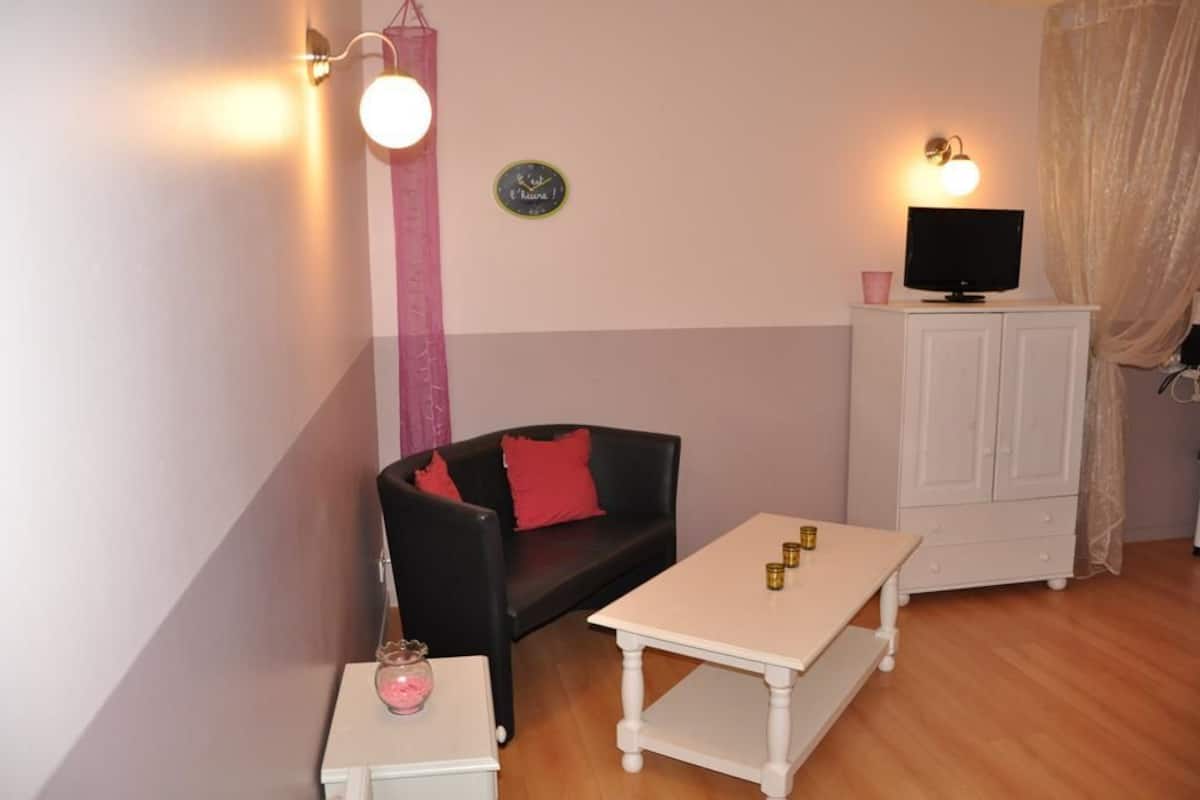A small room with a black sofa, a white table, a television on a cabinet, and a wall-mounted light fixture.
