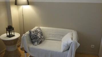 A white sofa with a geometric-patterned pillow, a small round table with a lamp, and a tall floor lamp in a neutral-colored room.