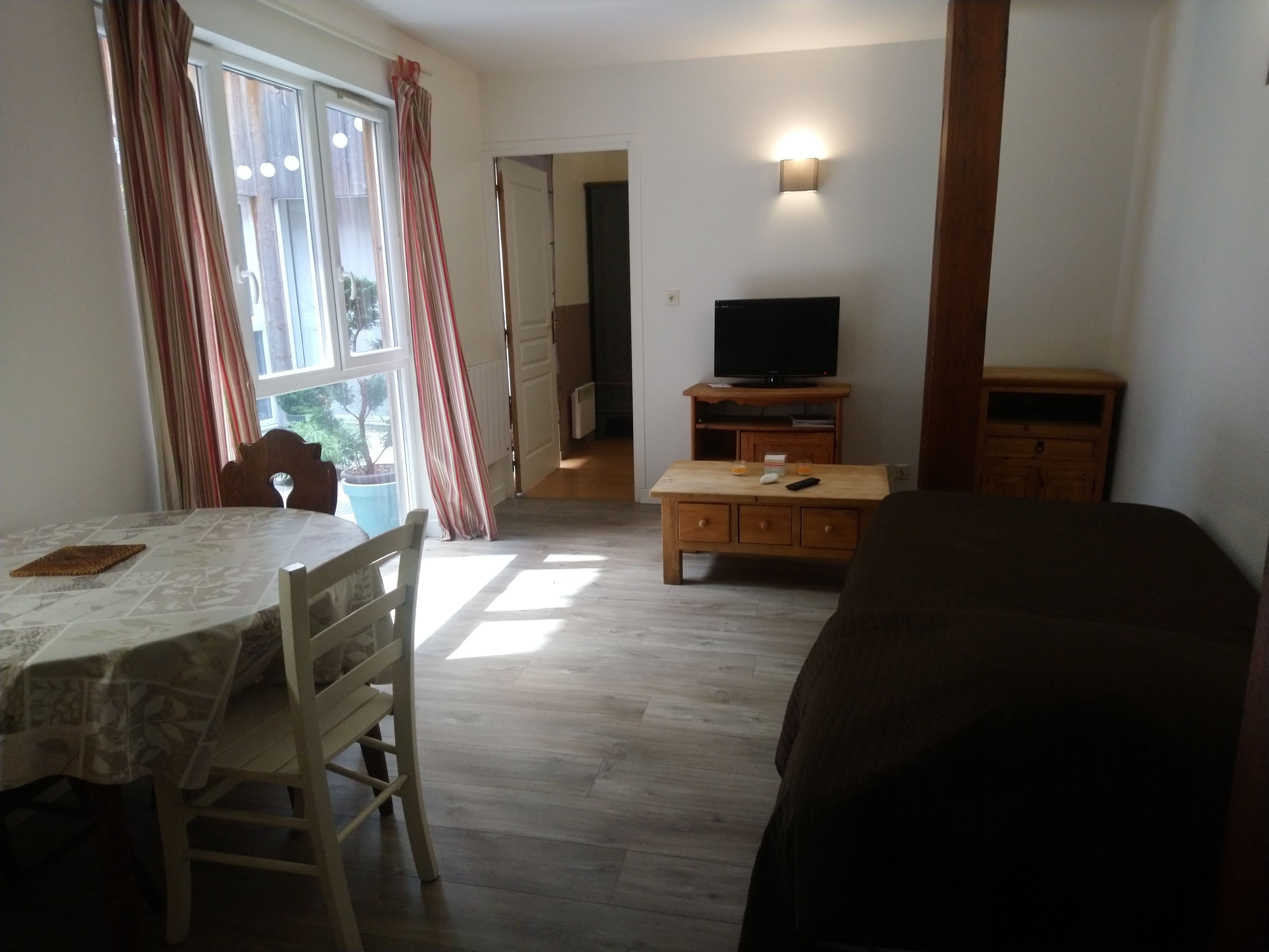 A room with a dining table, chairs, a TV on a wooden stand, a sofa, and a window with curtains.