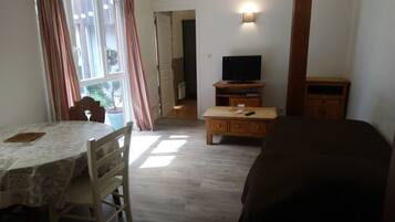 Una habitación con una mesa de comedor, sillas, un televisor sobre un soporte de madera, un sofá y una ventana con cortinas.