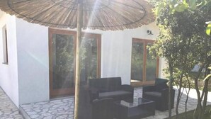 An outdoor seating area with a thatched umbrella, stone flooring, and a modern building with large windows. - Holiday home Sintija (Rab)