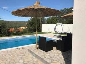 Une piscine aux bords recouverts de pierres, un parasol en chaume et des meubles d’extérieur noirs.