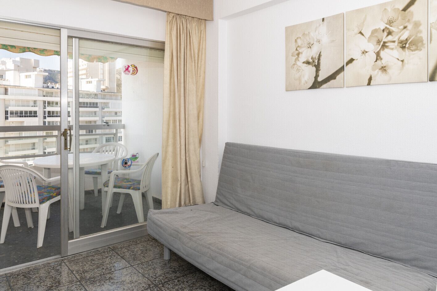 A room with a grey sofa, a glass door leading to an outdoor area with a table and chairs, and a beige curtain.