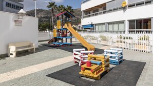A playground with a slide and climbing structures, a bench, and a building with balconies and a satellite dish. - PRADO, A-7º-72 PK INT 83 (Cullera)