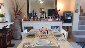 A dining area with a wooden table set for four, surrounded by white chairs. A large vase with branches and flowers is on the table, and there are various kitchen items and a television on the counter. - After8 B&B (Kleinbaai)