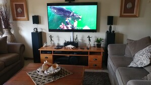 A living room with a TV mounted on the wall, a wooden entertainment unit with various items, a coffee table with decorative shells, and a sofa. - After8 B&B (Kleinbaai)