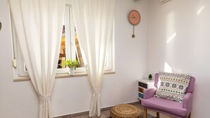 Una habitació amb finestra, cortines, un rellotge, una cadira morada, un pouf de fibra i una petita planta al davant de la finestra.