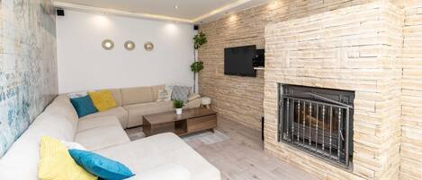 A modern living room with a stone fireplace, a large beige sofa with colorful cushions, a flat-screen TV mounted on the wall, and a small side table with a plant.