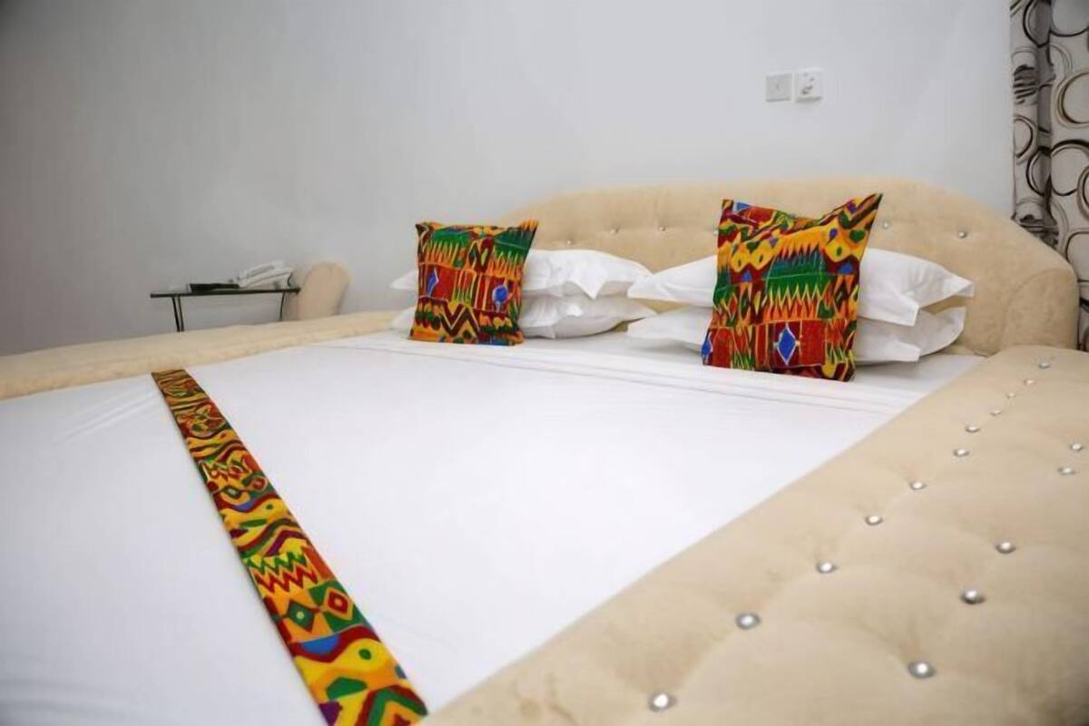 Una cama con sábana blanca, dos almohadas decorativas, un cabecero capitonado y una mesita de noche con un teléfono y una lámpara.