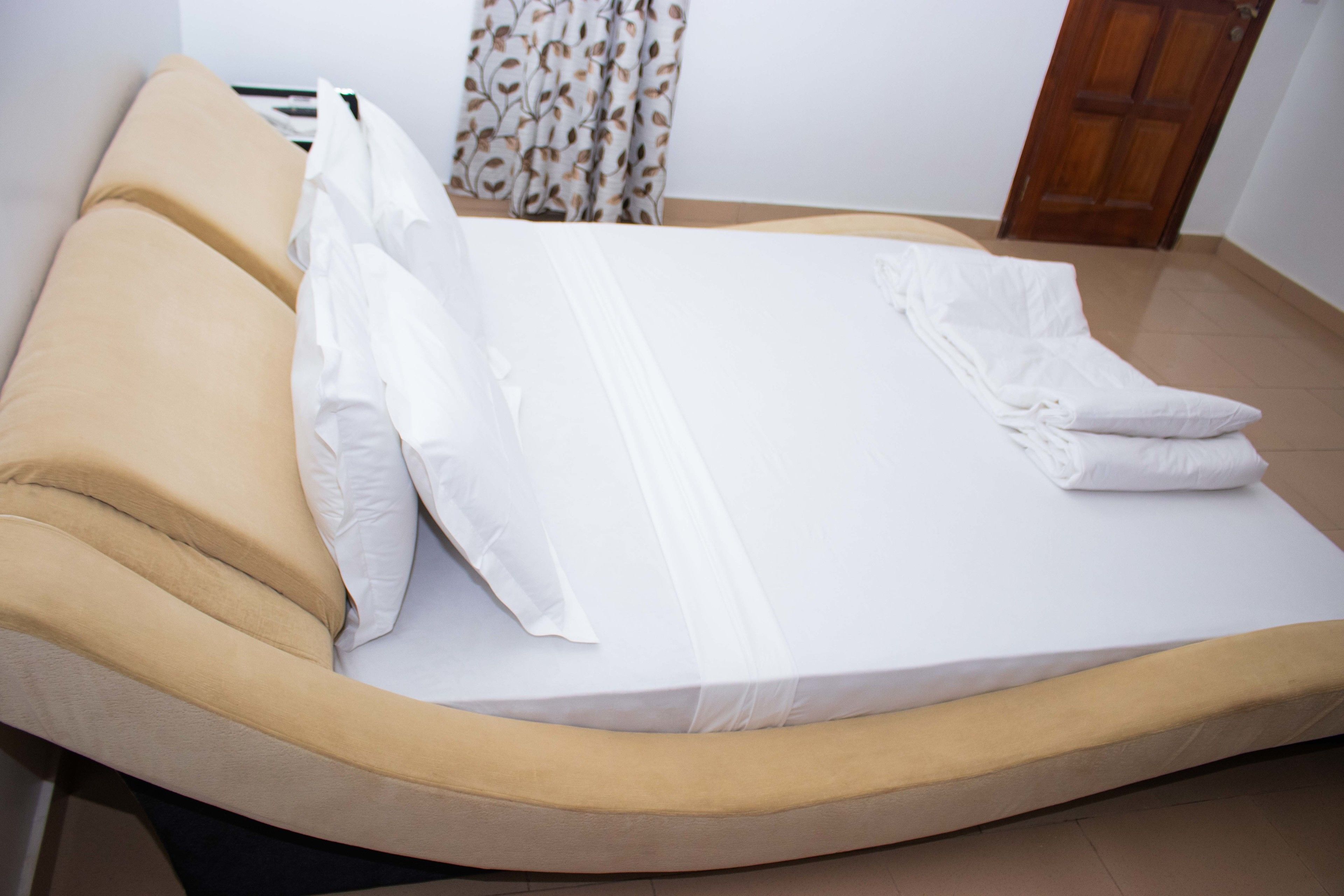 Un lit bien fait, avec du linge de lit blanc et une tête de lit beige, dans une pièce dotée d’une porte en bois et de rideaux à motifs.