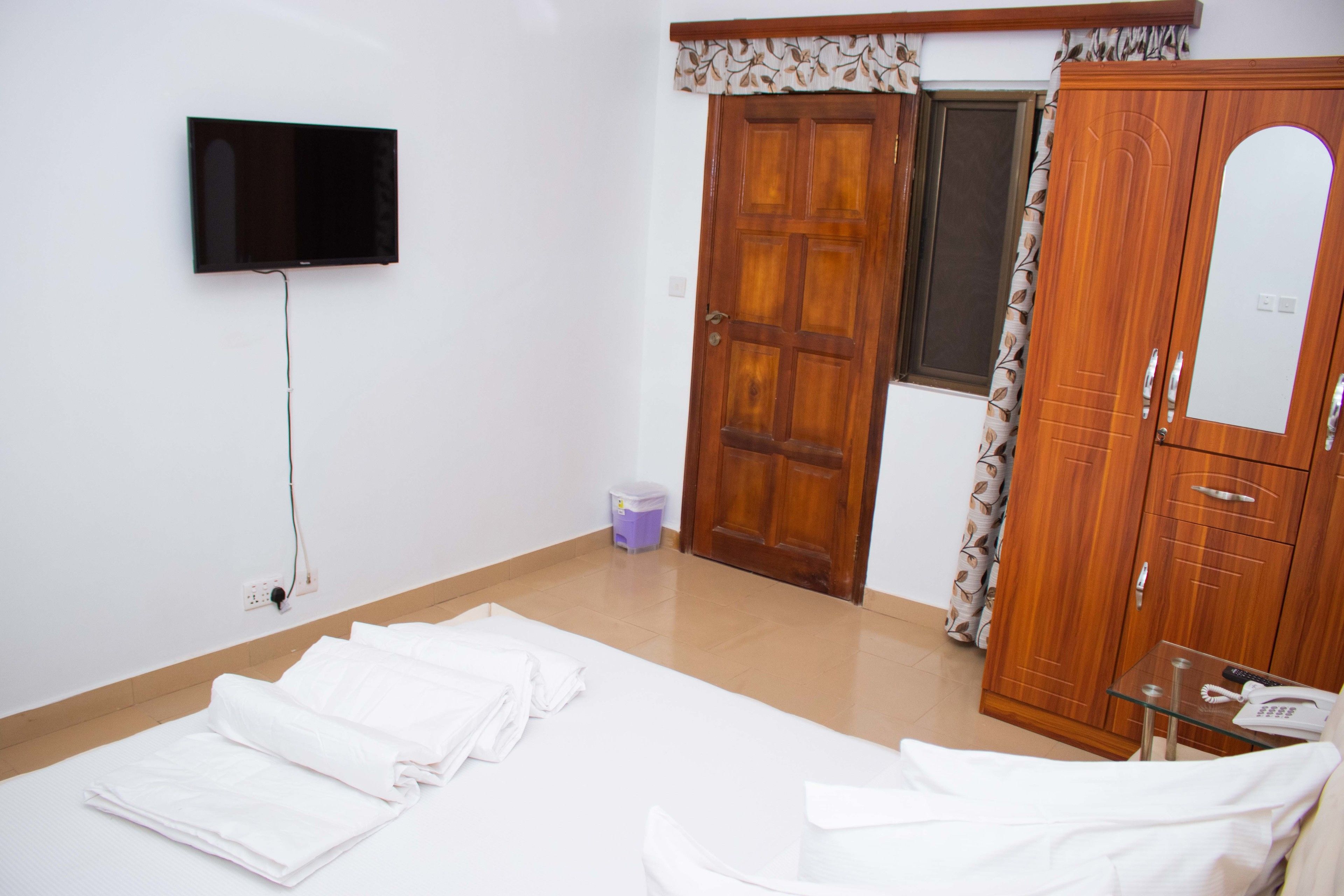 A hotel room with a bed, a TV mounted on the wall, a wooden wardrobe, and a small table with a phone and remote control.