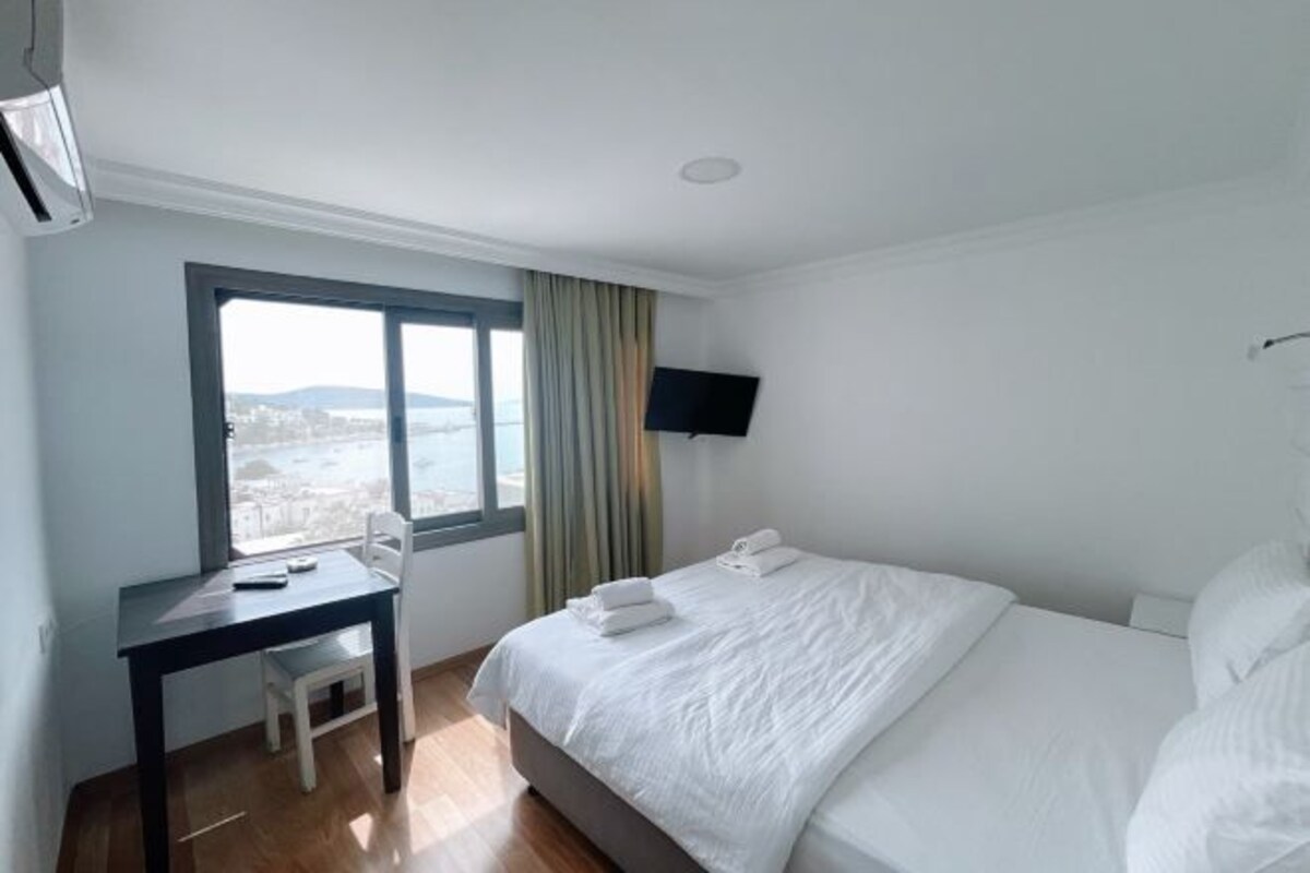Ett hotellrum med en säng, ett skrivbord, en stol, en tv som är monterad på väggen och ett fönster med utsikt över staden.