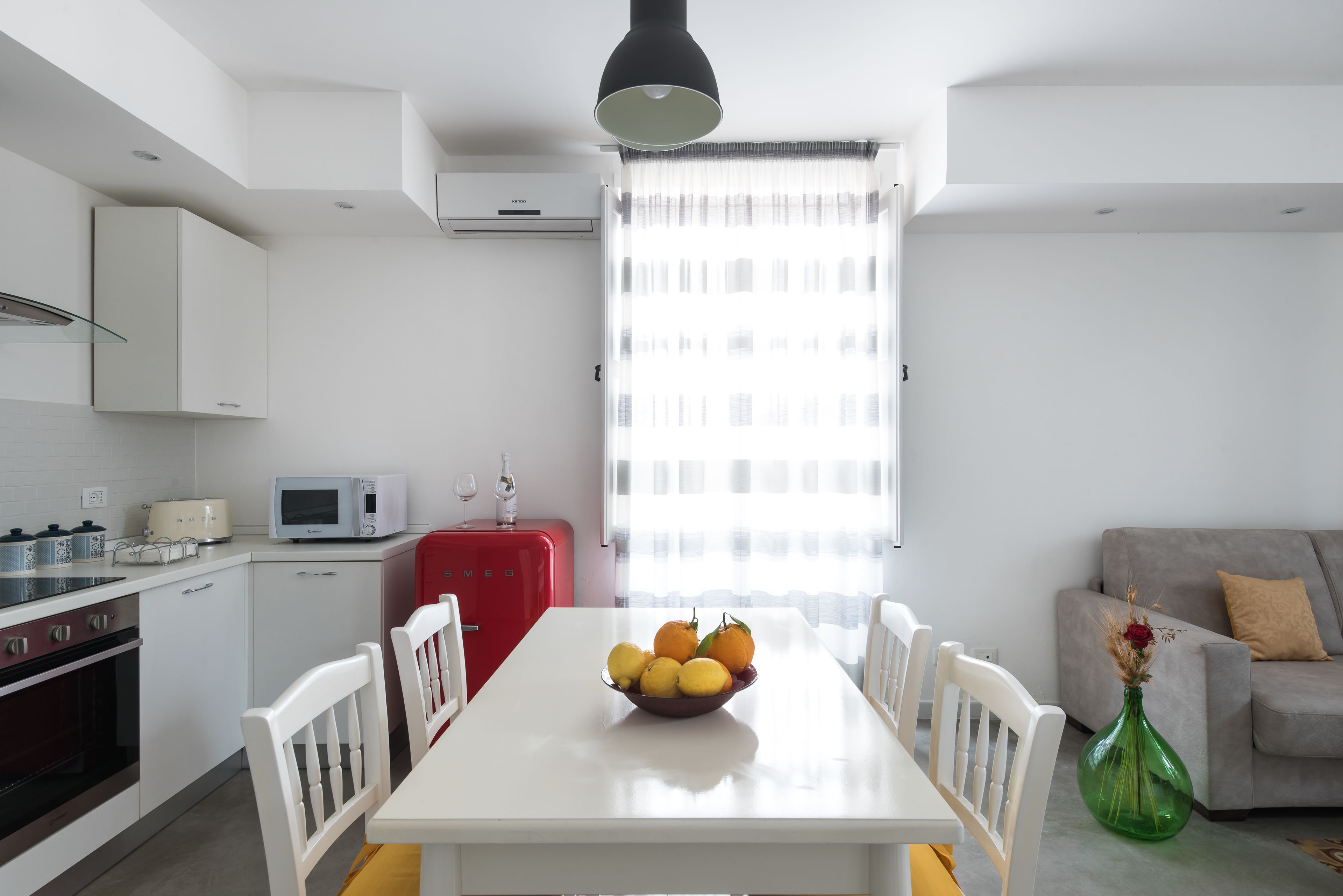 A modern kitchen with white cabinets, a red refrigerator, and a dining table with a bowl of fruit.